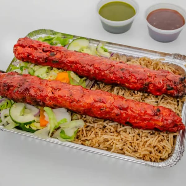 Two red chicken seekh kebabs on rice and salad in a foil container with two sauces.