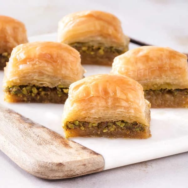 Five pieces of golden-brown baklava with pistachio filling on a white and wood serving board.